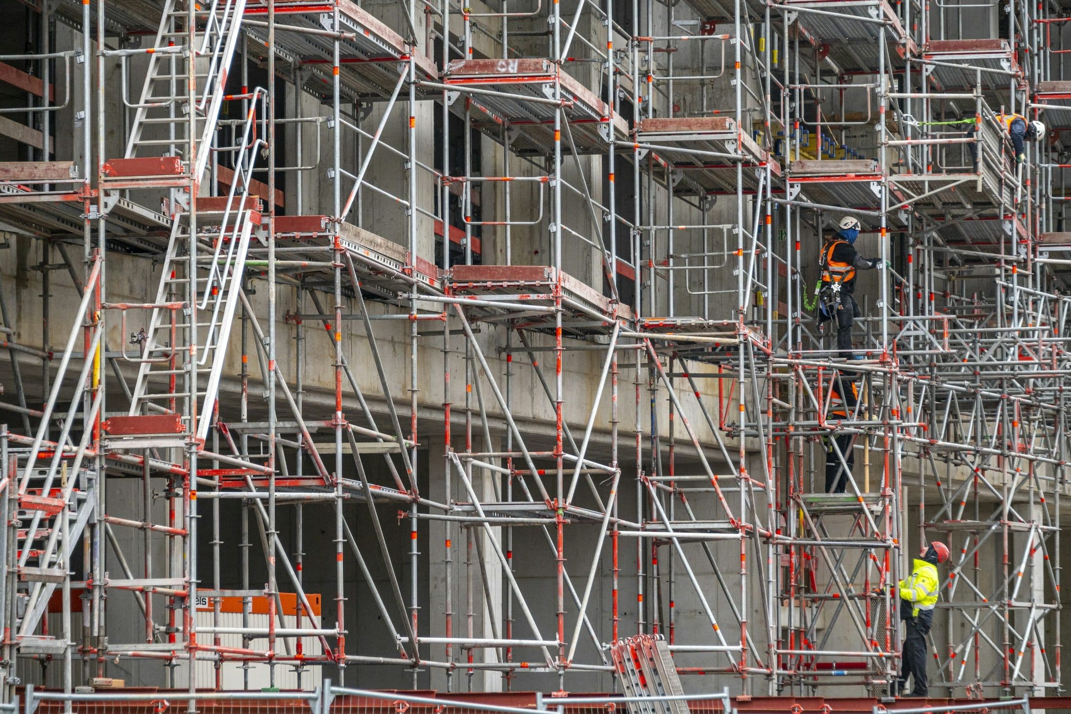 Un immeuble en béton en cours de construction, ceinturé d'échafaudages, avec des ouvriers en gilet haute visibilité sur les plateaux